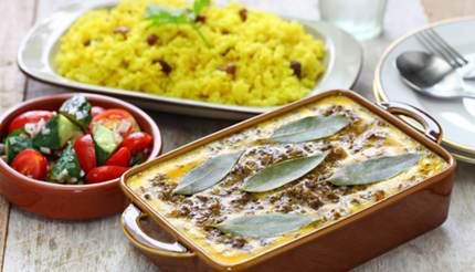 Bobotie and yellow rice in separate dishes, cucumbers and tomatoes in a bowl on the side