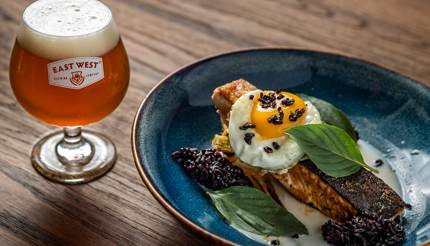 A protein-packed meal. Salmon on a blue plate with a fried egg on top and a glass of East West beer on the left side